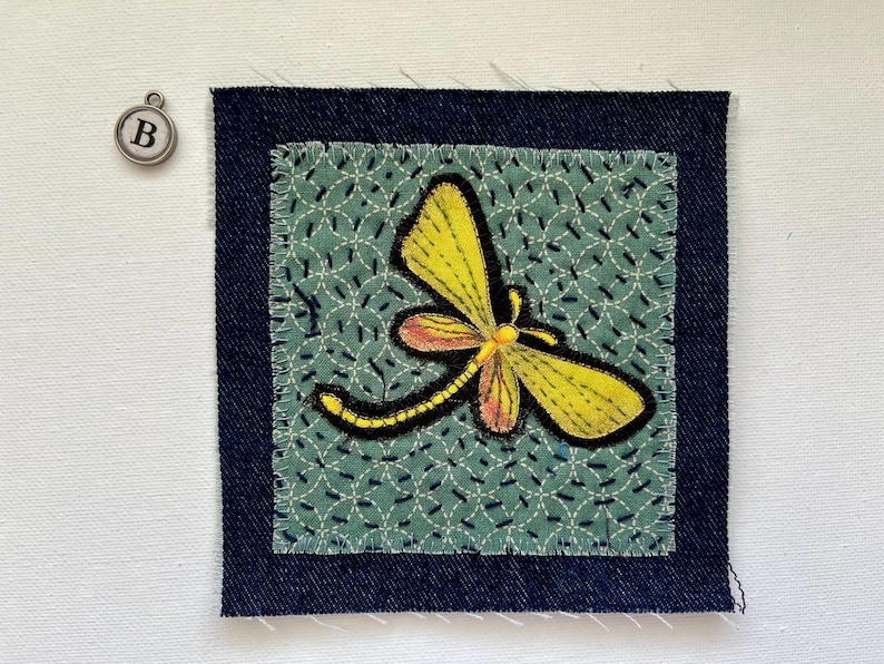 May include: A square fabric patch with a yellow and orange dragonfly design, stitched onto a teal background with a white pattern. The patch is framed by a dark blue denim border. A silver charm with the letter "B" hangs nearby.