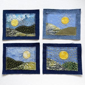 May include: Four rectangular fabric patches featuring embroidered landscape scenes. Each patch displays a yellow sun, blue sky, and green hills crafted from various patterned textiles, all framed by a dark blue denim border with zigzag stitching.