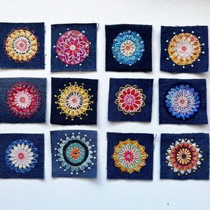 May include: Twelve square patches of dark blue denim fabric, each featuring a unique, hand-embroidered circular mandala design in vibrant, multi-colored thread. The embroidery patterns include various floral and geometric shapes in shades of yellow, red, pink, blue, and green, arranged in a grid of three rows and four columns.