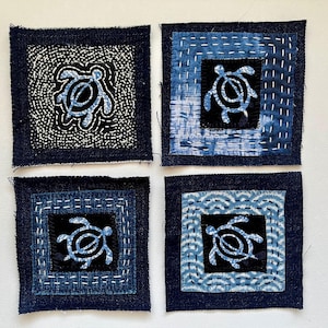 May include: Four square fabric patches with a dark blue denim border. Each patch features a blue turtle design on a black background. The turtles are surrounded by various patterns and stitching, creating a handcrafted aesthetic.