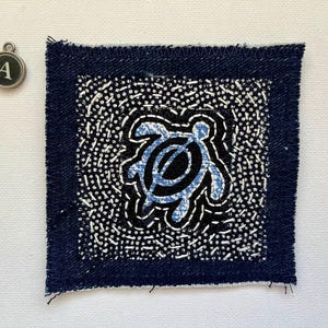 Peut inclure: Un patch de tissu bleu marine carr&eacute; avec un bord effiloch&eacute;. Il pr&eacute;sente un motif central de tortue bleu clair sur fond noir, entour&eacute; d'un motif de points blancs. Un charm argent&eacute; avec la lettre "A" est &eacute;galement visible.