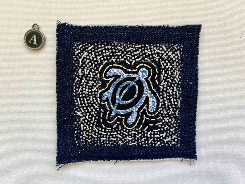 Peut inclure: Un patch de tissu bleu marine carr&eacute; avec un bord effiloch&eacute;. Il pr&eacute;sente un motif central de tortue bleu clair sur fond noir, entour&eacute; d'un motif de points blancs. Un charm argent&eacute; avec la lettre "A" est &eacute;galement visible.