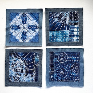 May include: Four indigo blue and white textile squares with various stitched patterns. Each square is framed with denim fabric. The designs include geometric shapes, radiating lines, and circular patterns, showcasing a handcrafted aesthetic.