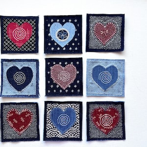 May include: Nine square fabric patches arranged in a grid, each featuring a heart shape with white stitched details. The hearts are in shades of red, blue, and light blue, set against dark blue backgrounds with various white patterns like dots and waves.