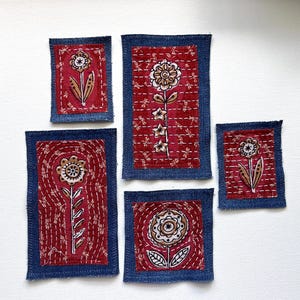 May include: Five rectangular fabric patches featuring red floral designs on a dark blue denim border. Each patch displays a hand-stitched flower motif with white thread detailing, showcasing a repeating dragonfly pattern on the red fabric sections.