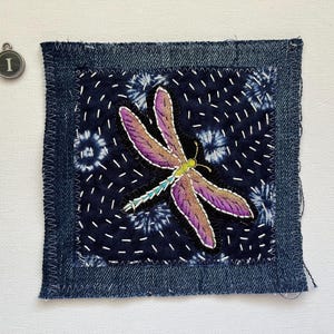 May include: A square textile patch with a denim border and a dark blue center featuring a dragonfly design. The dragonfly has purple and brown wings, a green body, and white stitching. An 'I' charm is in the upper left corner.