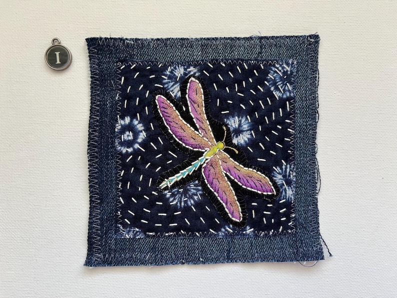 May include: A square textile patch with a denim border and a dark blue center featuring a dragonfly design. The dragonfly has purple and brown wings, a green body, and white stitching. An 'I' charm is in the upper left corner.