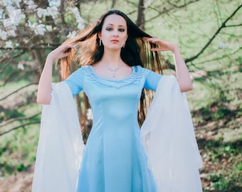 May include: A woman wearing a light blue gown with a white sheer overlay stands in a garden setting. She has long dark hair and is wearing a silver necklace.