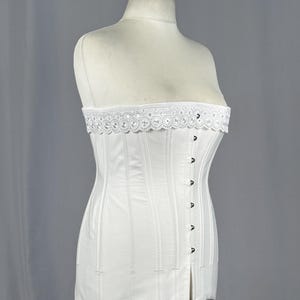 May include: A white strapless corset featuring vertical paneling and a front closure with metal hooks. The top edge is finished with a decorative white eyelet lace trim. The garment is displayed on a mannequin, showcasing its structured design.