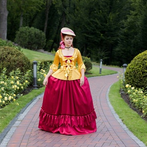 May include: A woman in a yellow and red dress with a large skirt and a hat stands on a brick pathway in a garden setting.