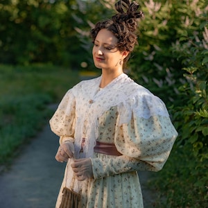 Puede incluir: Una mujer con un vestido de estilo vintage con estampado floral y ribete de encaje. Está de pie en un entorno de jardín con follaje verde y flores.