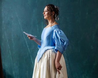 May include: A woman in a blue and white dress stands in front of a teal green wall. She is holding a piece of paper in her hand and looking to the side.