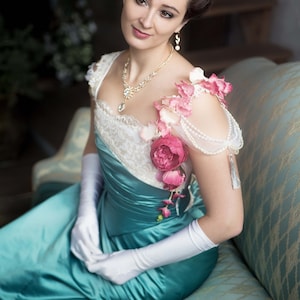 May include: A woman in a teal satin dress with a white lace bodice and a floral embellishment on the shoulder. She is wearing white gloves and a tiara.