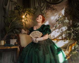 Puede incluir: Un vestido largo de color verde esmeralda con mangas abullonadas voluminosas, un escote amplio y una falda plisada completa. La prenda se combina con un delicado abanico plegable de color crema con recortes de encaje y guantes blancos.