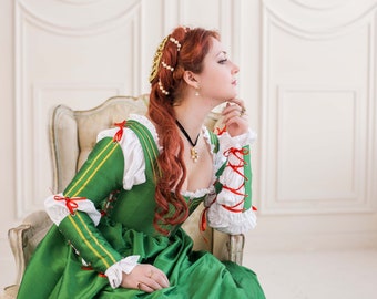 May include: A woman in a green and white dress with red accents sits in a white chair. She has long red hair and is wearing a gold crown.