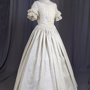 May include: A vintage cream-colored dress with a floral pattern and short ruffled sleeves. The dress features a fitted bodice and a full skirt, displayed on a mannequin. The fabric has a subtle striped texture.