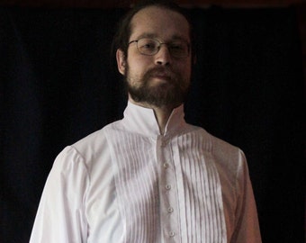 May include: A white dress shirt with a high collar and pleated front. The shirt has a button-up closure and long sleeves. The sleeves have a gathered cuff. The shirt is tucked into dark blue pants with a button closure.