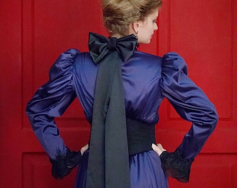 May include: A woman in a purple satin dress with a large black bow at the back of the neck. The dress has long sleeves and a fitted waist. She is standing in front of a red door.