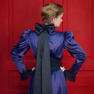 May include: A woman in a purple satin dress with a large black bow at the back of the neck. The dress has long sleeves and a fitted waist. She is standing in front of a red door.