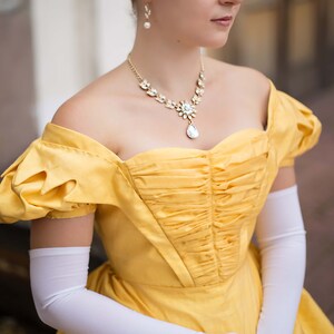May include: A woman wearing a yellow off-the-shoulder dress with a ruffled bodice and white gloves. She is wearing a delicate necklace with a teardrop pendant.