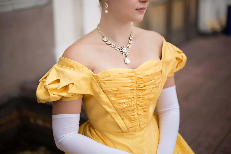 May include: A woman wearing a yellow off-the-shoulder dress with a ruffled bodice and white gloves. She is wearing a delicate necklace with a teardrop pendant.