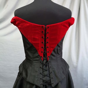 Op de afbeelding: Zwarte en rode corset met een veter aan de achterkant. De corset heeft een sweetheart-halslijn en een getailleerd lijfje. De achterkant van de corset is met zwart lint vastgezet.