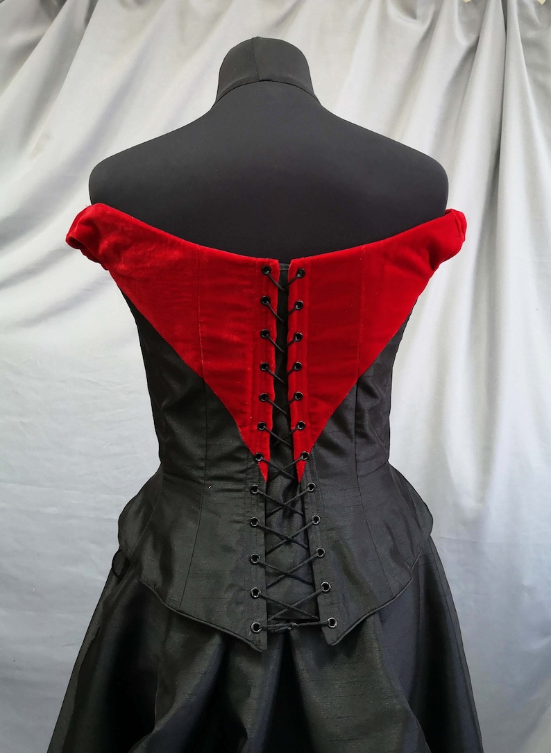 Op de afbeelding: Zwarte en rode corset met een veter aan de achterkant. De corset heeft een sweetheart-halslijn en een getailleerd lijfje. De achterkant van de corset is met zwart lint vastgezet.