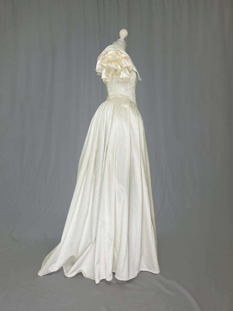 May include: A white satin wedding dress with a ruffled neckline and a full skirt.