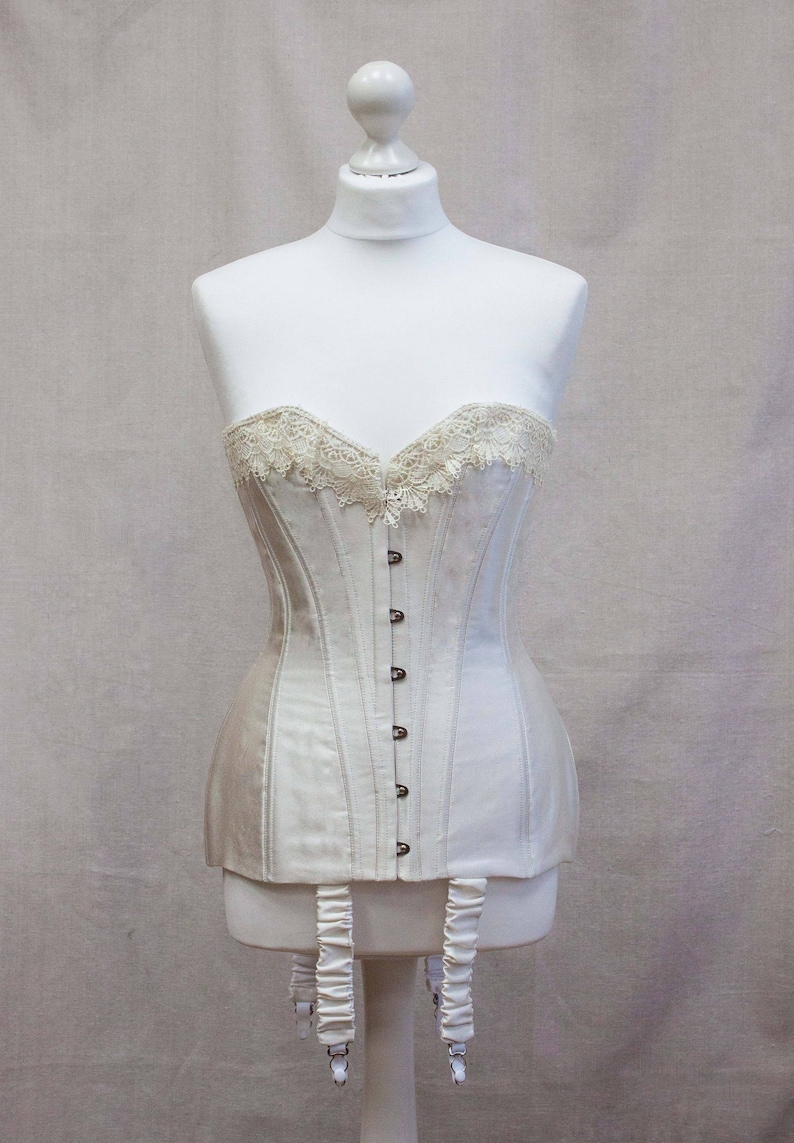 May include: A white strapless corset with lace trim and a row of buttons down the front. The corset has white garters attached to the bottom.