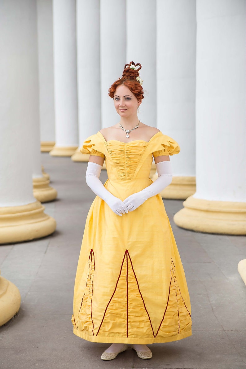 May include: A woman in a yellow gown with a red trim and white gloves stands in front of a row of white columns.
