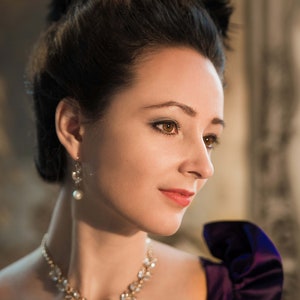 May include: A woman with dark hair styled in an updo wears a black dress with a purple satin bow on the shoulder. She is wearing a pearl earring and a diamond necklace with a teardrop pendant.