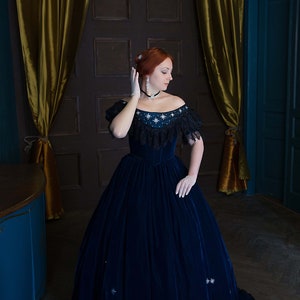May include: A woman wearing a blue velvet gown with a lace trim and star-shaped embellishments. She is standing in a room with gold curtains and a wooden door.