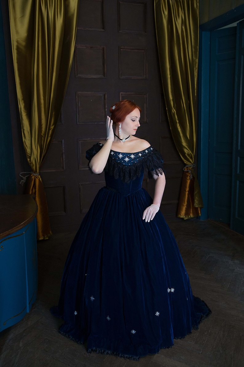 May include: A woman wearing a blue velvet gown with a lace trim and star-shaped embellishments. She is standing in a room with gold curtains and a wooden door.