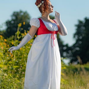 May include: A white Regency-style dress with a red bodice and white sleeves. The dress features a high waist and a flowing skirt. The model is wearing white gloves and a floral hairpiece. The scene is set outdoors in a field.