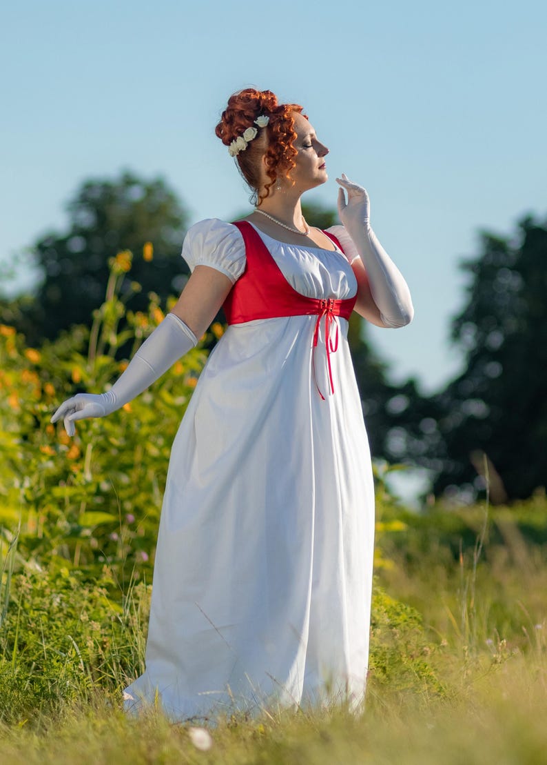 May include: A white Regency-style dress with a red bodice and white sleeves. The dress features a high waist and a flowing skirt. The model is wearing white gloves and a floral hairpiece. The scene is set outdoors in a field.