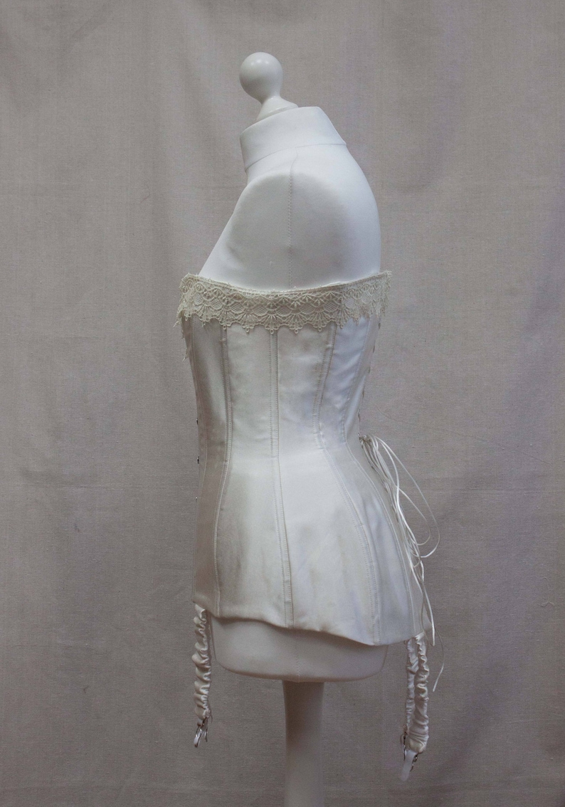 May include: A white satin corset with a lace trim and garter straps. The corset is on a white mannequin.