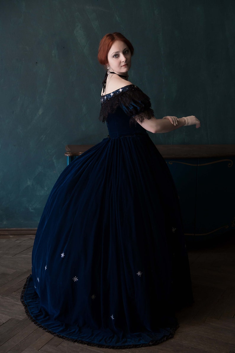 May include: A woman wearing a dark blue velvet gown with a lace overlay and silver star embellishments. She is standing in front of a dark green wall and a wooden cabinet.