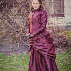 Op de afbeelding: Een vrouw in een bordeauxrode jurk met een rok met een bustle en een zwart jasje staat voor een bakstenen muur met wijnstokken. Ze draagt een zwarte hoed en handschoenen.