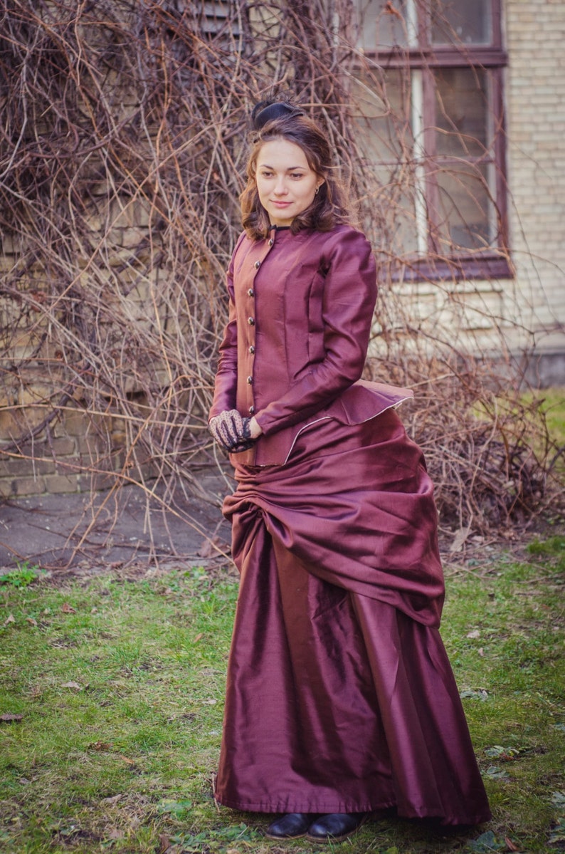 Op de afbeelding: Een vrouw in een bordeauxrode jurk met een rok met een bustle en een zwart jasje staat voor een bakstenen muur met wijnstokken. Ze draagt een zwarte hoed en handschoenen.