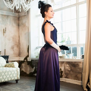 May include: A woman in a long, dark purple dress with a white lace glove and a black feather hat stands in a room with a large window and a chandelier.