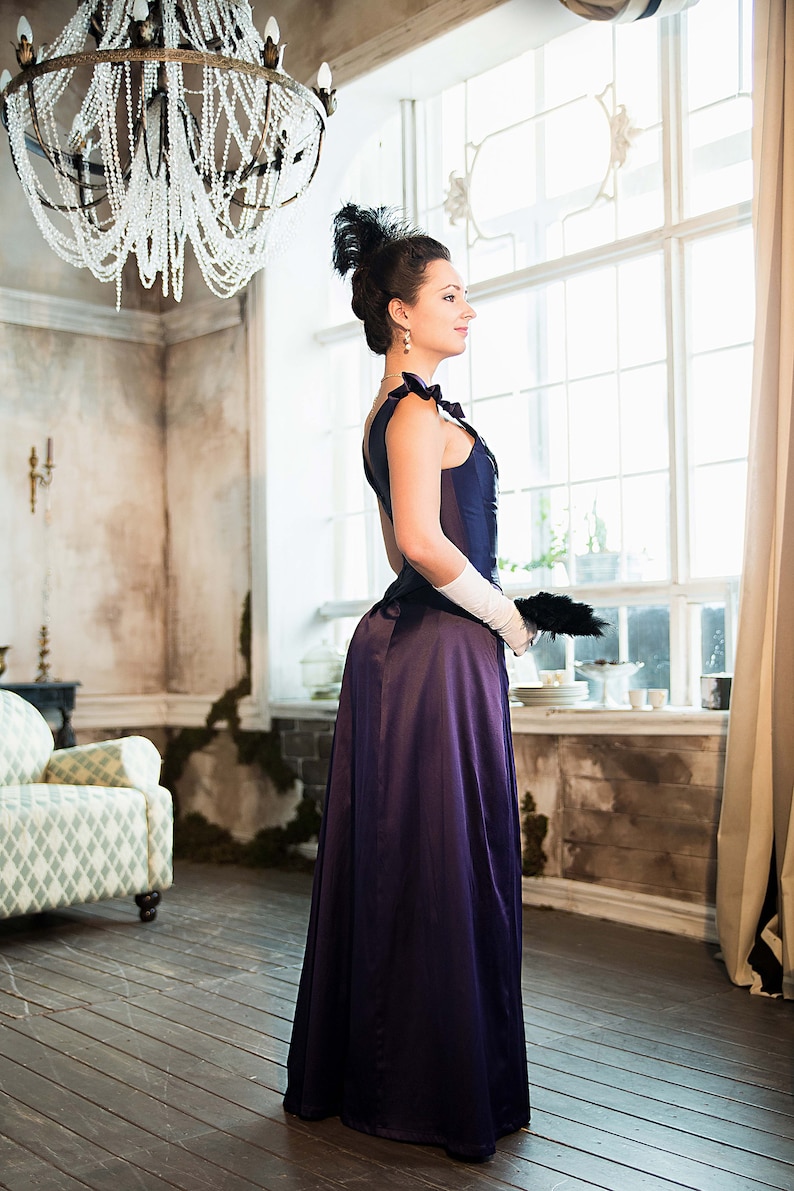 May include: A woman in a long, dark purple dress with a white lace glove and a black feather hat stands in a room with a large window and a chandelier.
