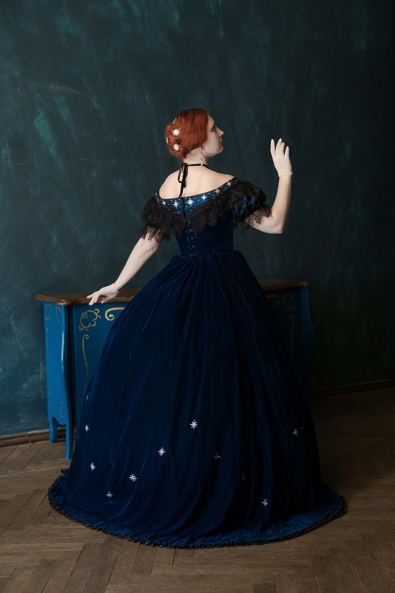 May include: A woman wearing a dark blue velvet gown with a black lace trim and white star embellishments. The gown has a full skirt and a fitted bodice. The woman is standing in front of a dark green wall and a blue chest of drawers.