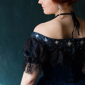 May include: A woman wearing a blue velvet dress with a black lace overlay and silver star embellishments. The dress has an off-the-shoulder neckline and a fitted bodice.