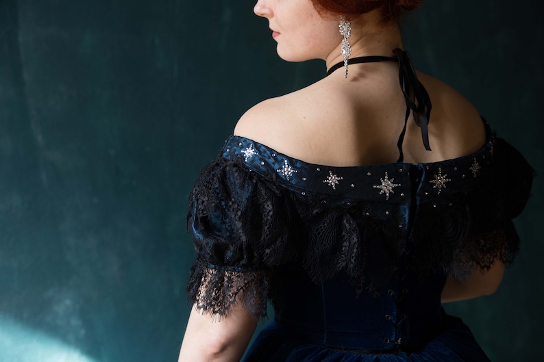 May include: A woman wearing a blue velvet dress with a black lace overlay and silver star embellishments. The dress has an off-the-shoulder neckline and a fitted bodice.