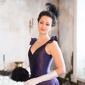 May include: A woman in a purple dress with a black lace bodice and a black feather in her hair. She is holding a black feather fan and wearing white gloves.