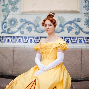 May include: A woman in a yellow gown with white gloves and a pearl necklace stands in front of a tiled wall.