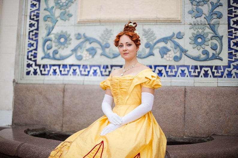 May include: A woman in a yellow gown with white gloves and a pearl necklace stands in front of a tiled wall.