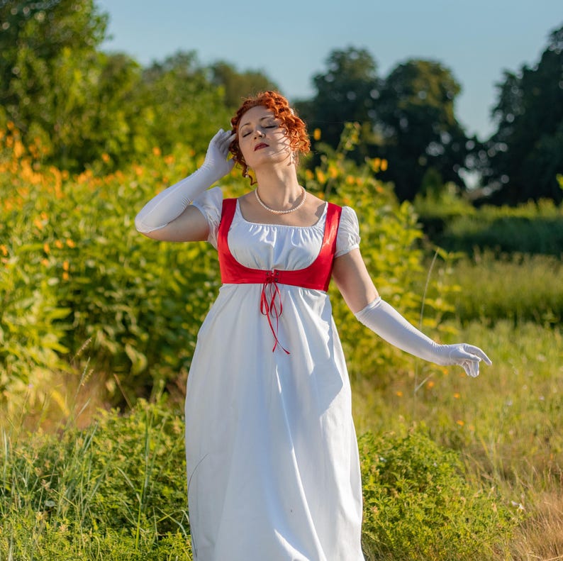 May include: A white Regency-style dress with a red bodice and red ribbon ties. The dress has short sleeves and is paired with long white gloves. The woman in the image is wearing a pearl necklace and has red hair.