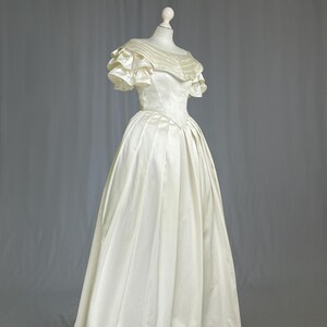 May include: A white satin wedding dress with a ruffled neckline and short sleeves. The dress has a fitted bodice and a full skirt.