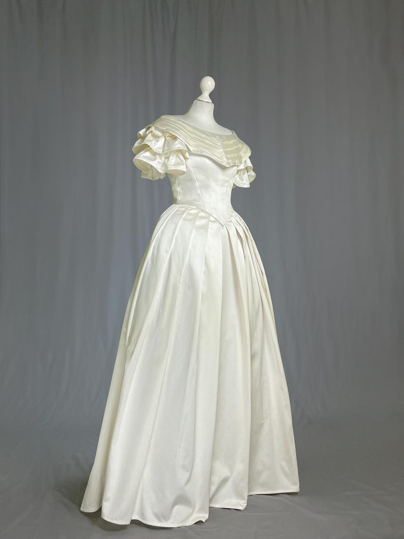 May include: A white satin wedding dress with a ruffled neckline and short sleeves. The dress has a fitted bodice and a full skirt.